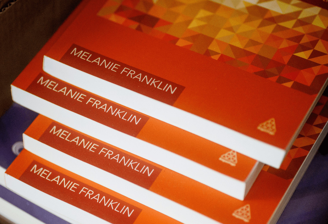 Stack of four orange marketing booklets with the authors name, Melanie Franklin, clearly visible which illustrates the continued importance of print media in the debate of print vs digital marketing.