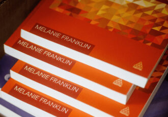 Stack of four orange marketing booklets with the authors name, Melanie Franklin, clearly visible which illustrates the continued importance of print media in the debate of print vs digital marketing.